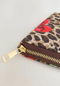Pochette en tissu à motif léopard avec un design de fleur rouge et une fermeture éclair en métal doré sur un bord en tissu marron, posée sur une surface claire.