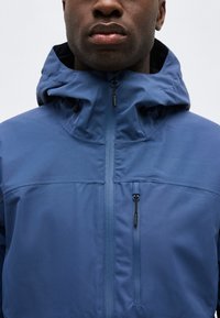 Homme portant une veste à capuche bleue avec une fermeture éclair frontale et une poche poitrine zippée, sur un fond clair uni.