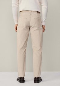 Personne portant un pantalon casual beige et des chaussures en cuir brun foncé, debout de dos face à l'appareil photo sur un fond uni.