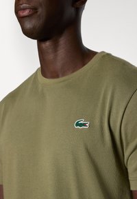 Ελιές πράσινη βαμβακερή μπλούζα με λαιμόκοψη και μικρό κεντημένο λογότυπο κροκόδειλου στο αριστερό στήθος. Λείο υφασμά.