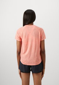 Γυναίκα με μακριά μαλλιά που φοράει ένα κοραλί κοντομάνικο μπλουζάκι και μαύρα αθλητικά σορτς, στέκεται μπροστά σε ένα απλό ανοιχτό φόντο.