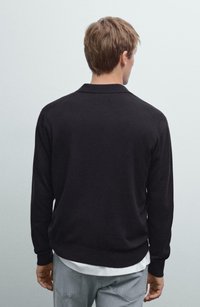 Man with light brown hair wearing a black long-sleeve shirt and light blue jeans, facing away against a plain light background.