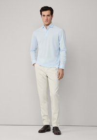 Svijetloplavi dukserica s dugim rukavima i malom kragnom, u kombinaciji s kremastim hlačama i tamnim loafericama. Glatka tekstura, ležeran dizajn.