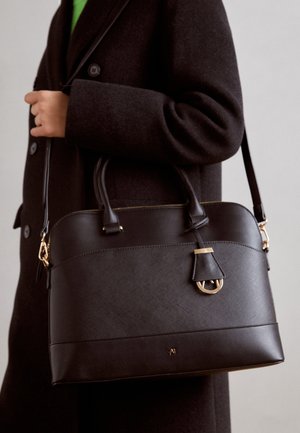 Persoon met een donkere jas die een grote gestructureerde donkerbruine leren handtas met handvatten en gouden accenten vasthoudt.