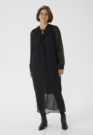 Femme souriante portant une longue robe noire à manches transparentes, décolleté à nouer, leggings noirs et bottines noires, sur fond blanc.