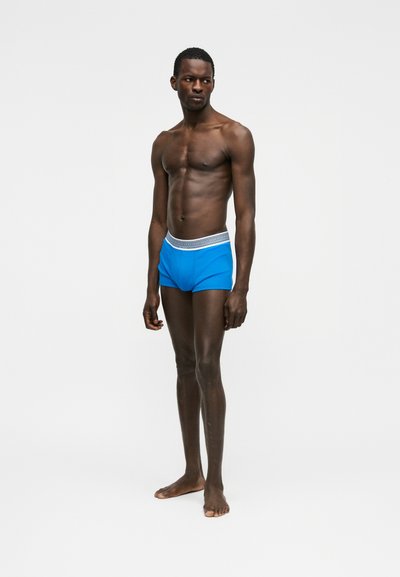 Modèle masculin à la peau foncée, debout pieds nus, portant un boxer bleu avec une ceinture à motifs, sur un fond blanc uni.