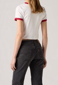 Camiseta blanca corta con cuello y puños acanalados rojos, combinada con jeans gris oscuro que presentan un diseño distintivo en el bolsillo trasero y una etiqueta de logo.
