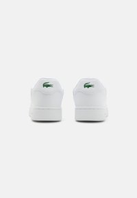 Hvite sneakers med grønn Lacoste krokodillelogo på hælen, glatt lær overdel, rund tå og flat gummisåle.