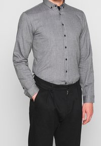 Camicia grigia con bottoni, realizzata in un tessuto strutturato, colletto a tab e bottoni neri, abbinata a pantaloni neri plissettati.