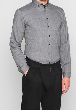 Chemise grise à boutons avec un tissu texturé, col à pattes et boutons noirs, associée à un pantalon plissé noir.