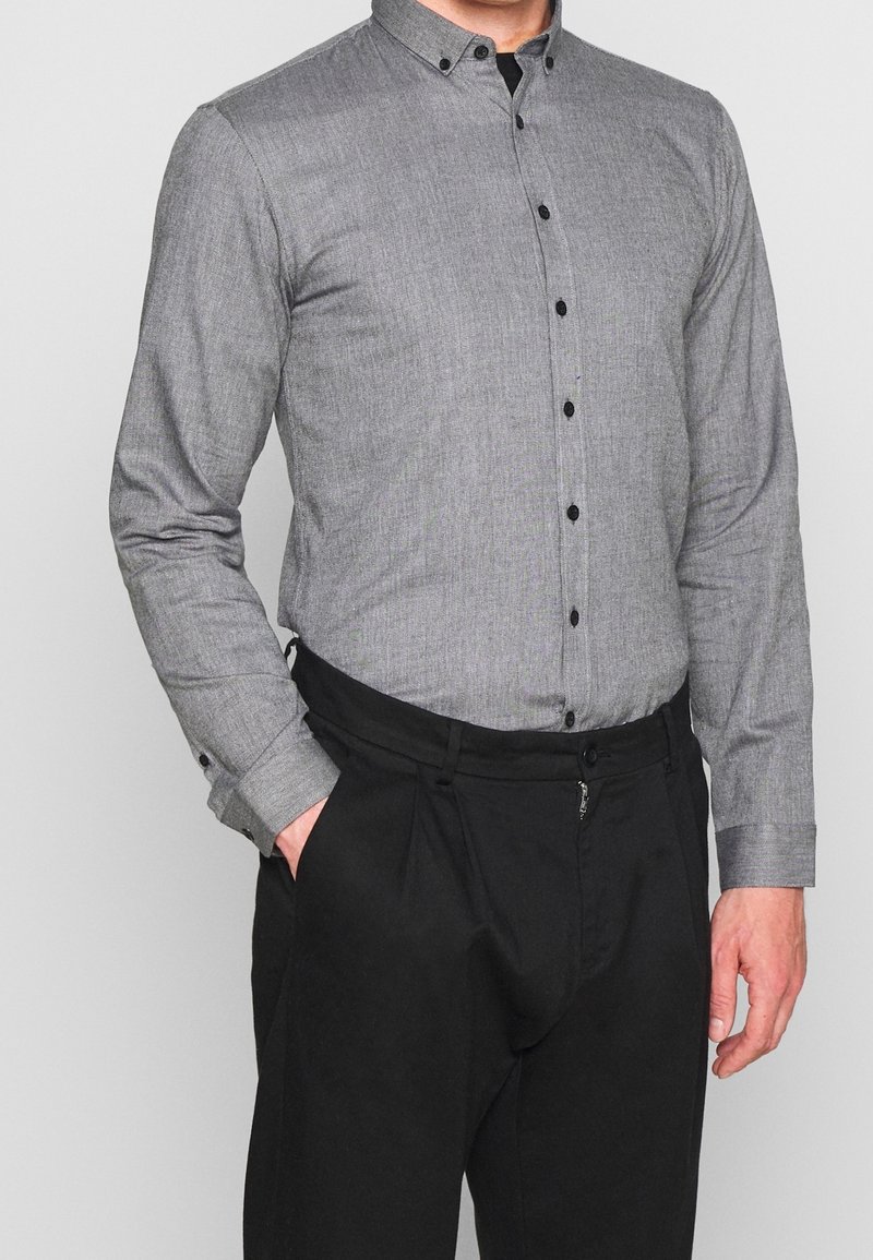 Camicia grigia con bottoni, realizzata in un tessuto strutturato, colletto a tab e bottoni neri, abbinata a pantaloni neri plissettati.