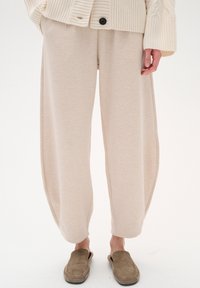 Pantaloni a gamba larga beige in morbido misto cotone con una vestibilità rilassata. Presentano lati drappeggiati e una superficie liscia. Abbinati a scarpe marroni slip-on.