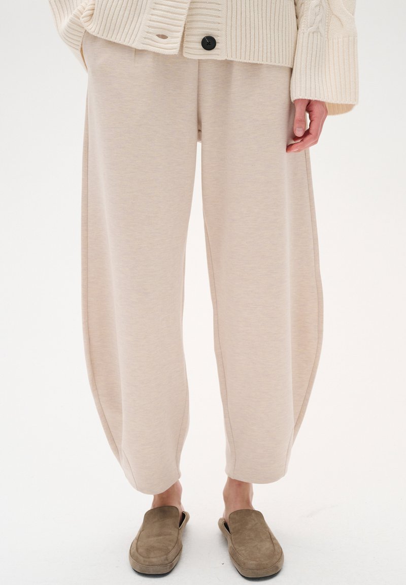 Pantaloni a gamba larga beige in morbido misto cotone con una vestibilità rilassata. Presentano lati drappeggiati e una superficie liscia. Abbinati a scarpe marroni slip-on.