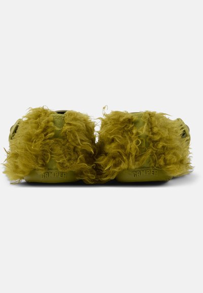 Pair of olive green slippers with thick curly fur covering and "Camper" embossed on the back sole, shown from the rear.