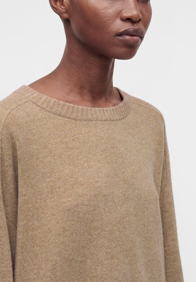 Gros plan d'une personne portant un pull en maille beige à col rond sur un fond clair uni.