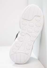 Svart sportskor med en strukturerad vit gummisula, med cirkulära greppmönster och varumärkeslogotyp på insidan.