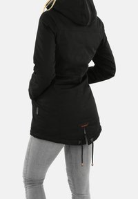 Parka noire à capuche avec une taille cintrée, des cordons de serrage au bas et deux poches latérales. Présente une étiquette de logo brun à l'arrière.