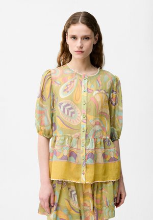 Femme aux longs cheveux ondulés portant un chemisier à motifs pastel avec des manches bouffantes et une jupe assortie sur un fond blanc uni.