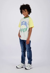 Vaikas su garbanotais plaukais stovi šypsodamasis, dėvi geltonomis rankovėmis marškinėlį „Social Surfer“, mėlynas džinsus ir baltus sportbačius, priešais paprastą foną.