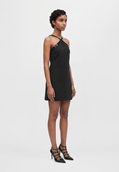 Un mannequin aux cheveux courts et bouclés porte une mini-robe noire à bretelles croisées et des chaussures noires à talons avec des brides. Il se tient devant un fond clair et uni.
