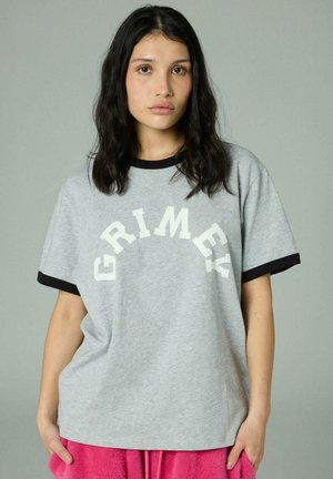 Mujer con cabello largo y oscuro que lleva una camiseta gris con borde negro y texto blanco "GRIMEY", combinada con pantalones rosa intenso, posando frente a un fondo gris.