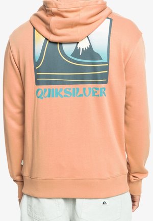 Persona que lleva una sudadera con capucha color melocotón con un gráfico azul y negro y texto "QUIKSILVER" en la espalda, combinada con pantalones de color claro.