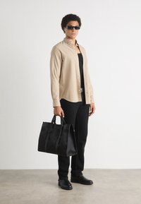 Camicia beige a bottoni sopra una canotta nera, abbinata a pantaloni e scarpe neri. Tiene in mano una grande borsa a tracolla nera con texture.