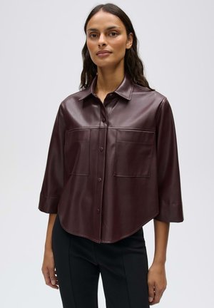Vrouw draagt een donkerbruine leren blouse met knopen, driekwart mouwen en twee borstzakken, gecombineerd met een zwarte broek.