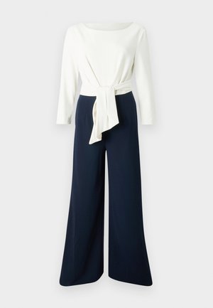 Haut blanc à manches longues avec un détail de taille nouée, associé à un pantalon large bleu marine, présentant un tissu lisse et une silhouette décontractée.
