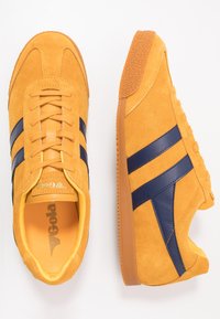 Sneaker gialle in suede con strisce blu navy, suola in gomma texturizzata e lacci abbinati. Presentano un colletto imbottito e un logo sulla linguetta.