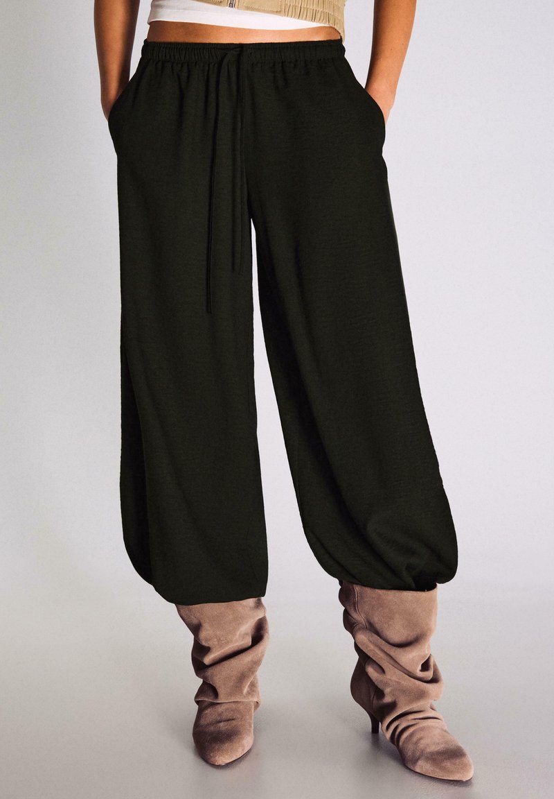 Femeie purtând pantaloni negri largi cu mansete elastice, asortați cu cizme cu toc din piele intoarsa taupe, lejere, cu mâinile în buzunare.