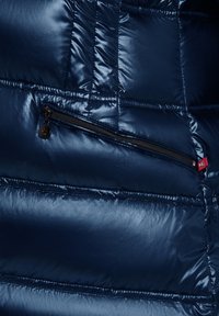 Marineblauwe, glanzende pufferjackstof met gestikte delen. Beschikt over een ritszak met een zwarte rits en een rode trekstrip.