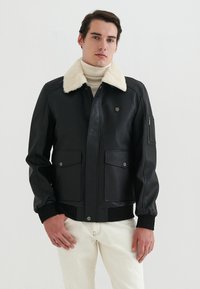 Svart läderjacka med en beige fuskpälskrage, framdragkedja, två stora bröstfickor och en ficka med dragkedja på ärmen.