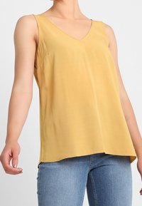 Donna che indossa un top giallo senza maniche con scollatura a V abbinato a jeans di denim blu, su uno sfondo chiaro e semplice.