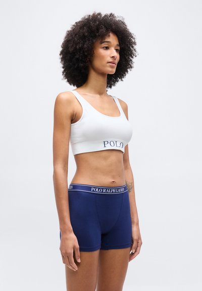 Γυναίκα με σγουρά μαλλιά, που φοράει λευκό αθλητικό σουτιέν και σκούρο μπλε σορτς με το λογότυπο Polo Ralph Lauren, στέκεται μπροστά σε απλό φόντο.