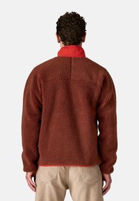 Uomo di spalle che indossa una giacca in pile marrone con un colletto rosso e pantaloni beige su uno sfondo chiaro e uniforme.