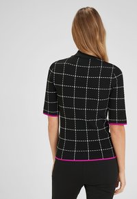 Žena s dlouhými vlasy, oblečená v černém topu s krátkým rukávem, který má bílou mřížkovanou vzor a růžové lemování na rukávech a spodním lemu, viděná zezadu.