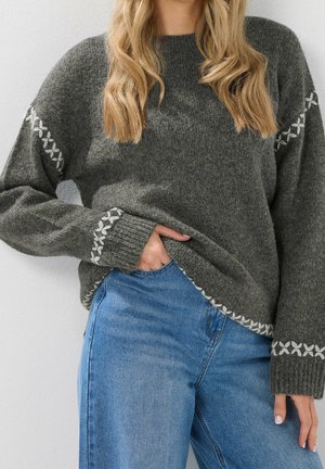 Femme portant un pull en tricot gris avec des motifs blancs cousus sur les manches et l'ourlet, associée à un jean bleu, debout avec une main dans la poche.