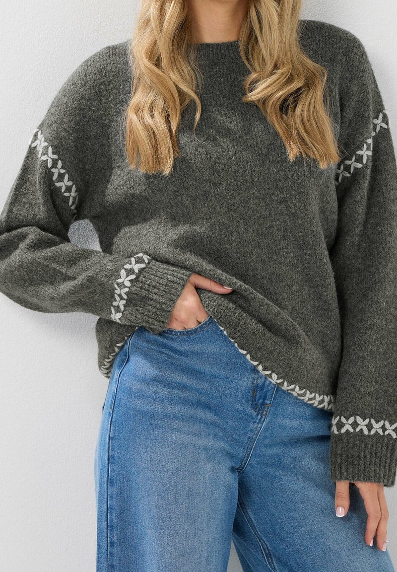 Femme portant un pull en tricot gris avec des motifs blancs cousus sur les manches et l'ourlet, associée à un jean bleu, debout avec une main dans la poche.