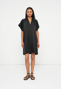 Vestido preto com decote em V, mangas curtas com babados e ajuste solto. Feito de tecido macio, chega à metade da coxa, combinado com sandálias pretas.