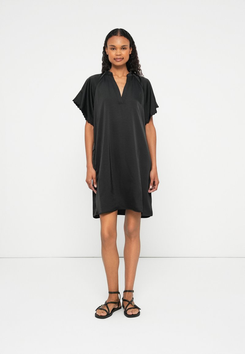 Črna obleka z V-izrezom, kratkimi nabranimi rokavčki in ohlapnim krojem. Narejena iz mehkega materiala, seže do sredine stegna, v kombinaciji s črnimi sandali.