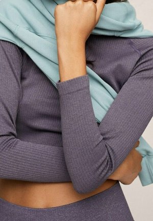 Femme portant un haut violet à côtes à manches longues et un bas assorti avec un vêtement bleu clair drapé sur ses épaules.