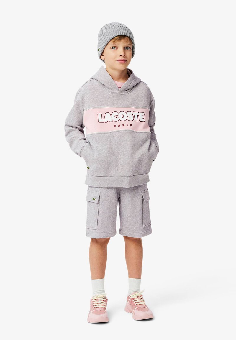 Felpa grigia con striscia rosa e scritta "LACOSTE PARIS", abbinata a pantaloni cargo grigi. Indossato con un berretto grigio e sneakers rosa.