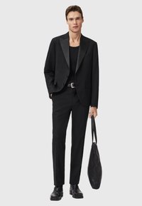 Completo nero con rever in satin, t-shirt nera sottostante, pantaloni su misura e stivali in pelle neri. Tiene una borsa a tracolla nera dalla texture particolare.