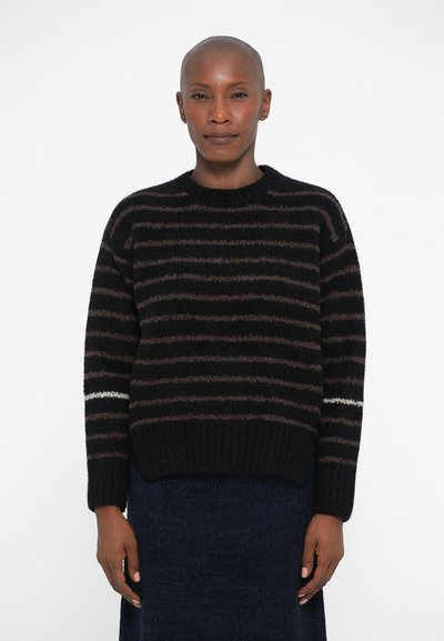 Person in a black and brown striped sweater with a white stripe on one sleeve, standing against a plain white background.
