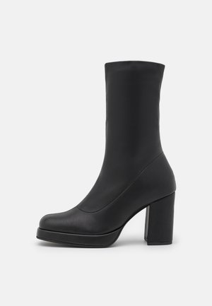 Botte en cuir noire mi-mollet avec un bout arrondi, une semelle plateforme et un talon haut épais, vue de côté sur un fond blanc.