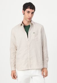 Light beige plaid shirt with green accent, featuring a button-up design, long sleeves, and a small embroidered logo on the chest.