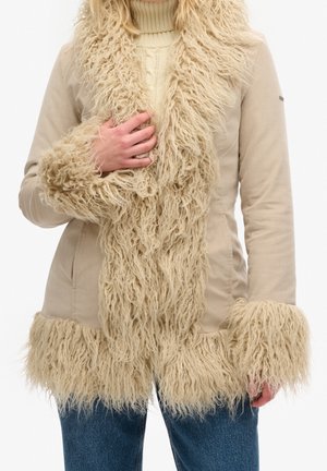 Femme portant un manteau beige avec des bordures en fourrure longue et hirsute sur le col, les poignets et l'ourlet, associé à un jean bleu et un pull en maille crème.