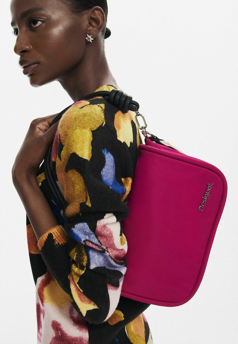 Borsa di tessuto rosa con forma rettangolare e superficie liscia, dotata di una tracolla nera e hardware in argento. Indossata sulla spalla contro un maglione colorato.