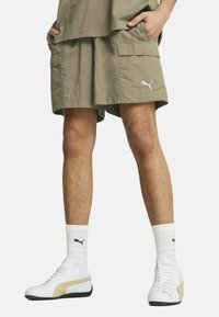 Maslinaste cargo kratke hlače s dva bočna džepa, širokog kroja, obične teksture i malim bijelim Puma logotipom. Uparene s bijelim i zlatnim tenisicama te prugastim čarapama.